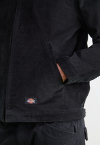 Osoba w czarnej kurtce z kordurowej tkaniny Dickies, z ręką w bocznej kieszeni, prezentująca zbliżenie tekstury kurtki oraz naszywki z logo.