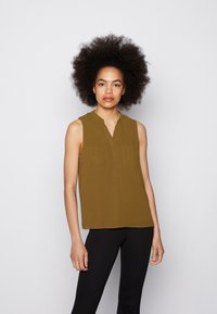 Blusa sem mangas verde-azeitona com colarinho, decote em V e dois bolsos no peito. Textura suave, corte ligeiramente solto, combinada com calças pretas.