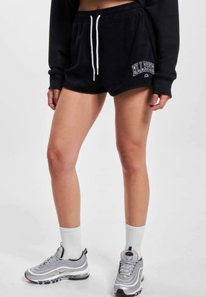 Persona vistiendo pantalones cortos negros de Ellesse con cordón blanco, medias blancas y zapatillas grises de Nike, de pie sobre un fondo liso.