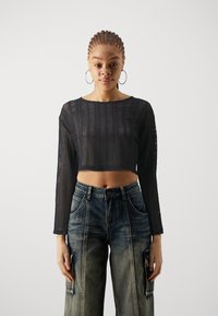 Black, cropped, long-sleeve top with a textured, open-knit design. Paired with high-waisted, faded blue denim cargo pants featuring patches.