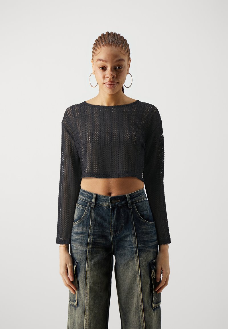 Black, cropped, long-sleeve top with a textured, open-knit design. Paired with high-waisted, faded blue denim cargo pants featuring patches.