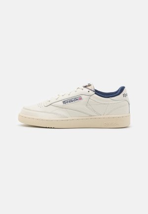 Baskets basses Reebok blanc cassé avec doublure intérieure bleu marine, bout perforé, panneaux cousus et logo de marque sur le côté et le talon.