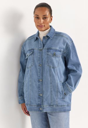 Frau steht und trägt eine hellblaue, übergroße Jeansjacke mit Knöpfen und Brusttaschen über einem weißen Rollkragenpullover.