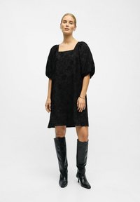 Vestido negro de mangas abullonadas con escote cuadrado, hecho de tela texturizada con un patrón floral, combinado con botas de cuero negro hasta la rodilla.