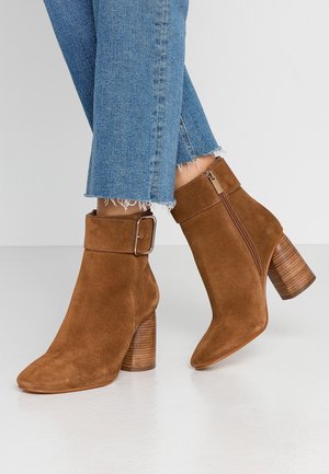 Bottines en daim brun avec un talon épais et texturé, fermeture éclair latérale, et large revers orné d'une grande boucle.