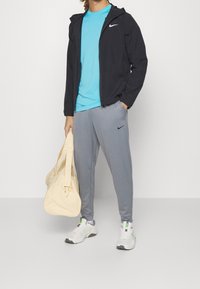 Man i svart zip-up jacka, blå skjorta, grå Nike sweatpants, vita sneakers, som håller en beige duffelväska med ena handen i fickan.