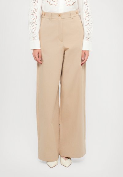Beige wide-leg trousers with a smooth texture, two front buttons, and side pockets, paired with a white patterned shirt and pointed shoes.