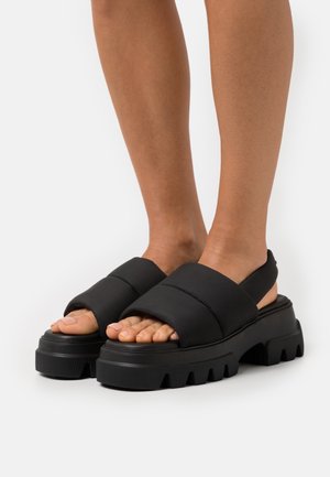 Sandales à plateforme noires avec des lanières épaisses matelassées, une semelle en caoutchouc texturée et un design à bout ouvert. Dotées d'une semelle épaisse à rainures pour une meilleure adhérence.