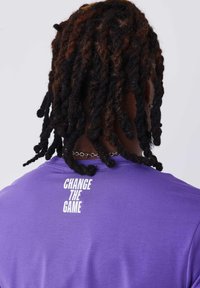 Camiseta de algodón morada de manga corta y cuello redondo, que presenta el texto "Cambia el juego" impreso en blanco en la parte trasera.