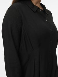 Ženska, ki nosi črno bluzo z dolgimi rokavi, ovratnikom in gumbi spredaj, prikazana od ramena do pasu.