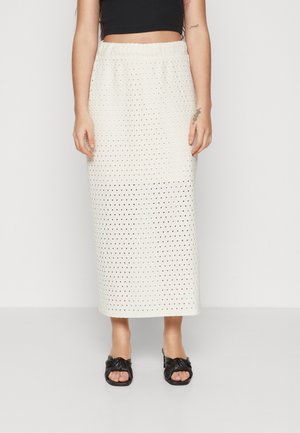 Vrouw draagt een witte midi-rok met perforaties, een zwart cropped top en zwarte knoop-sandalen, staand tegen een effen achtergrond.