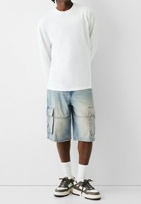 Bílá košile s dlouhým rukávem, světle modré cargo šortky se dvěma předními kapsami a tenisky v neutrálních tónech s tmavými akcenty a bílými tkaničkami.