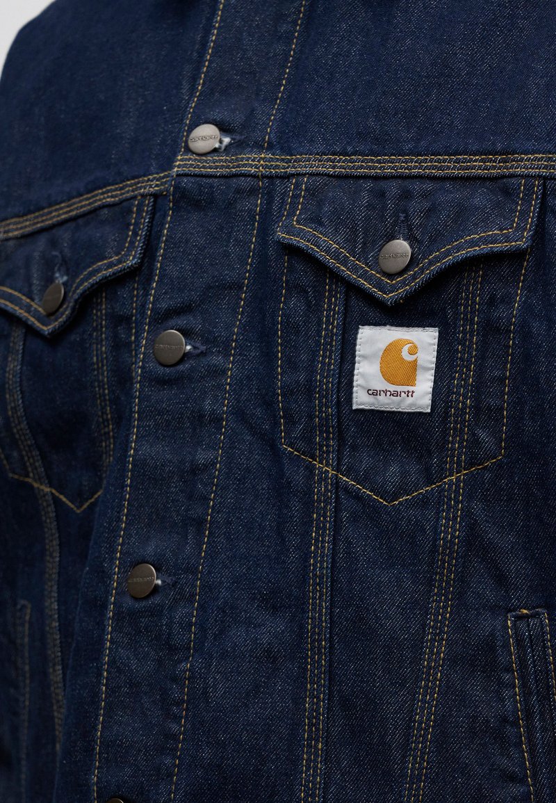 Dark blue denim jacket with yellow stitching, metal snap buttons, two chest pockets, and a white Carhartt logo patch on the right pocket.
