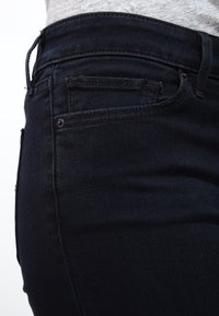 Primer plano de un bolsillo y la cintura de unos vaqueros de mezclilla azul oscuro, combinados con una camisa de color gris claro.