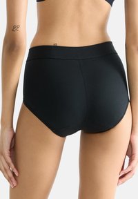 Svart högmidjad trosa med slät yta, elastiskt midjeband och vågiga kanter, designad för att sitta tajt och vara bekväm.