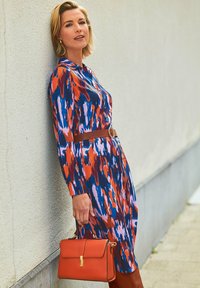 Multicolorkleurige blousejurk met een golvend patroon in blauw, rood en roze. Bruine ceintuur met een ronde sluiting en een oranje handtas.