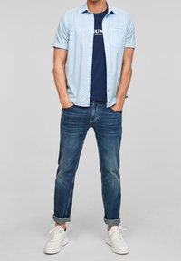 Camicia a maniche corte azzurro chiaro con piccoli motivi, indossata sopra una maglietta blu navy. Abbinata a jeans scuri arrotolati e sneakers bianche.