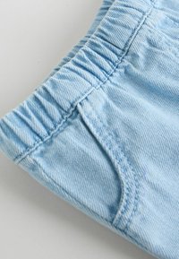 Lyseblå denimbukser med elastisk taljebånd og synlige syninger på forlommerne på en ensartet lys overflade.