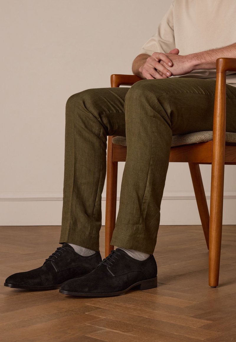 Zwarte suède dresschoenen met ronde neus en vetersluiting, gedragen met grijze sokken en olijfgroene broek, zittend op een houten stoel.