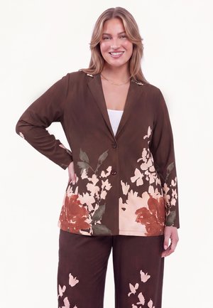 Smilende kvinde iført brun blazer med blomsterprint og matchende bukser, stående med den ene hånd på hoften mod hvid baggrund.