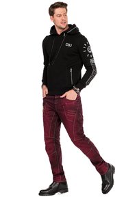 Zwarte hoodie met voorzakken met rits en logodetails, gecombineerd met bordeaux rode jeans met textuur en gestikte accenten en zwarte schoenen.
