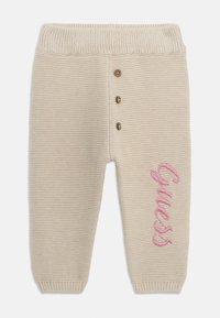 Beige Strickhose mit geripptem Bund, drei braunen Knöpfen an der Vorderseite und pinkem gesticktem "Guess" seitlich. Weiche Textur.