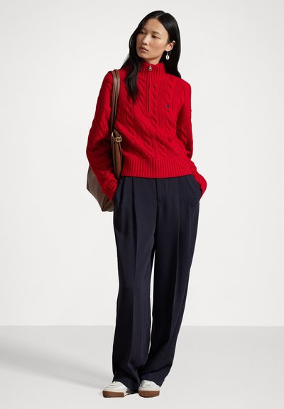 Red cable-knit sweater with a high collar, paired with navy wide-leg trousers. White sneakers and a brown bag complete the outfit.