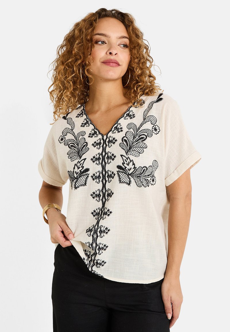 Blusa de color crema con intrincados bordados florales negros a lo largo del escote y las costuras de los hombros, con mangas cortas y un corte relajado.