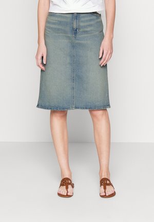 Knie-lange vervaagde blauwe spijkerrok gedragen met een wit shirt en bruine sandalen met open tenen tegen een lichte achtergrond.