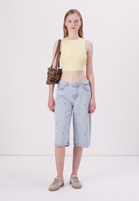 Un crop top côtelé jaune clair associé à des culottes en denim bleu clair ornées de broderies florales, complété par un sac à épaule marron et des baskets beiges.