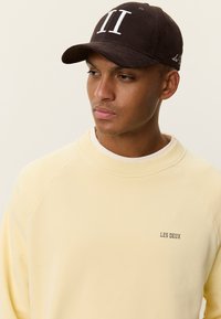 Καφέ καπέλο με λευκή κεντημένη επιγραφή "II", φορεμένο με απαλό κίτρινο φούτερ που διαθέτει μικρό λογότυπο "LES DEUX". Ομαλή υφή υφάσματος.