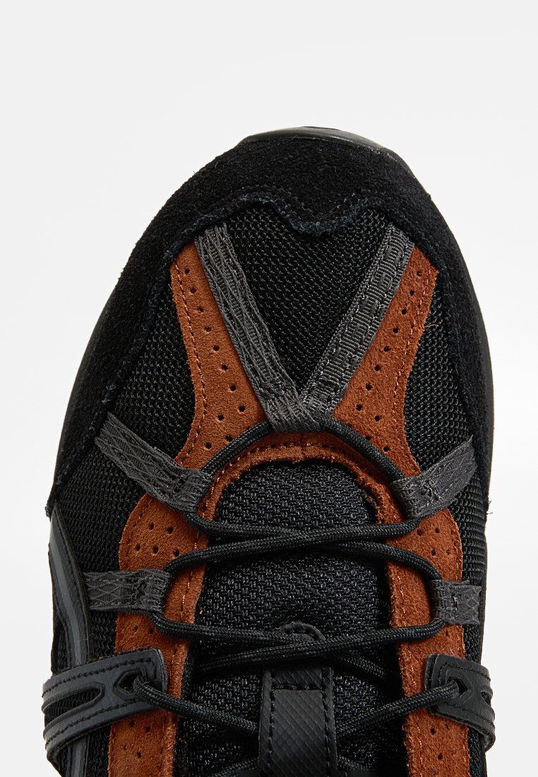Scarpa sportiva nera e marrone con materiali in rete e suede; presenta lacci grigi e una superficie testurizzata; punta arrotondata e occhielli rinforzati.