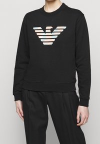 Felpa nera con collo tondo e logo a strisce colorate sul davanti. Tessuto morbido, con polsini e orlo a coste.