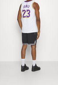 Weißes Basketballtrikot mit lila Akzenten, auf dem Rücken mit "JAMES" und der Nummer "23". Schwarze Shorts mit weißem Zickzacksaum und schwarze Sneakers.