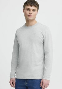 Light gray long-sleeve t-shirt made of a soft fabric, featuring a round neckline and subtle logo detail on the chest. Paired with blue jeans.