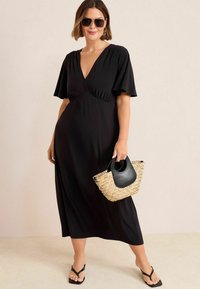 Robe maxi noire avec un décolleté en V profond, des manches volantées et une taille empire. Associée à un sac tissé beige avec une poignée noire et des sandales.