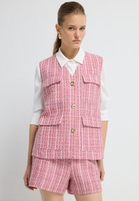 Růžová tkaninová vesta s vysokým límcem, čtyřmi kapsami a zlatými knoflíky, kombinovaná s odpovídajícími šortkami a bílou blůzkou s dlouhým rukávem.