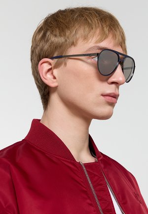Persona giovane con capelli corti castano chiaro, che indossa grandi occhiali da sole aviator scuri e una giacca rossa con cerniera, rivolta verso destra.