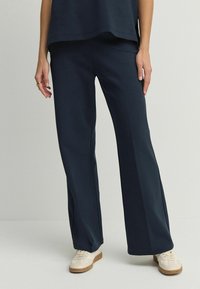 Pantaloni evazați bleumarin dintr-un material moale, având o textură netedă și o croială lejeră, asortați cu adidași de culoare deschisă.