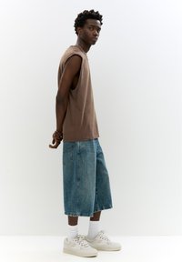 Camisetta marrone senza maniche, pantaloncini di jeans blu larghi e sneakers bianche con suole strutturate, in posizione eretta su uno sfondo bianco uniforme.