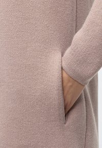 Main reposant dans la poche d'un pull tricoté doux rose clair avec un poignet visible sur la manche.