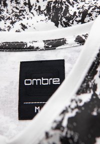 Černé a bílé tričko s texturovaným abstraktním vzorem. Výstřih je bílý s černou visačkou, na které je značka "ombre" a velikost "M".