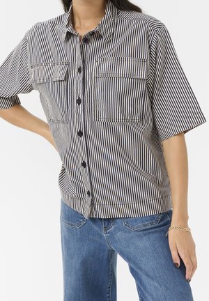 Personne portant une chemise à manches courtes rayée verticalement noire et blanche avec des poches poitrine et un jean bleu, debout avec une main sur la hanche.