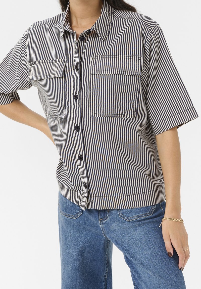 Personne portant une chemise à manches courtes rayée verticalement noire et blanche avec des poches poitrine et un jean bleu, debout avec une main sur la hanche.