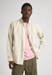 Light beige linen shirt with button-up design, worn over a pink T-shirt. Features long sleeves and a relaxed fit, with subtle texture.