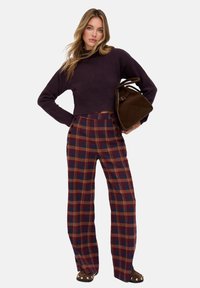 Pflaumenfarbener verkürzter Pullover, hoch taillierte karierte Hose in Rot, Marineblau und Beige mit Knopfdetails. Modell hält eine braune Tasche.