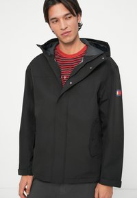 Άνδρας ντυμένος με μια μαύρη φούτερ ζακέτα πάνω από ένα κόκκινο και μαύρο ριγέ πουκάμισο, στέκεται μπροστά από ένα απλό ανοιχτό φόντο, κοιτάζοντας στο πλάι.