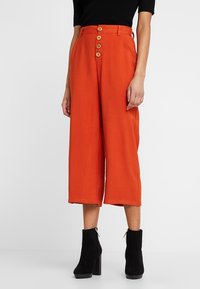Roodbruin gekorte broek met een losse pasvorm, voorzien van een vijfknopen sluiting aan de voorkant en een soepele stof, gecombineerd met zwarte enkelboots.