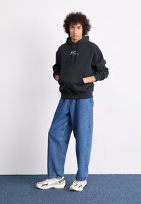 Crna dukserica s kapuljačom i logotipom, široke plave traperice i bijele tenisice s crnim detaljima. Model stoji naslonjen na jednostavnu pozadinu.