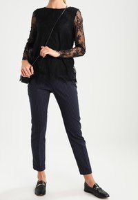 Top in pizzo nero con maniche lunghe trasparenti, design a strati, abbinato a pantaloni a righe blu navy e mocassini neri. Inclusa borsa a tracolla semplice.